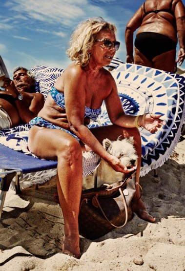 Mature woman in blue bikini sits with a small white dog on the beach, other beachgoers and blue sky in the background on a sunny day.