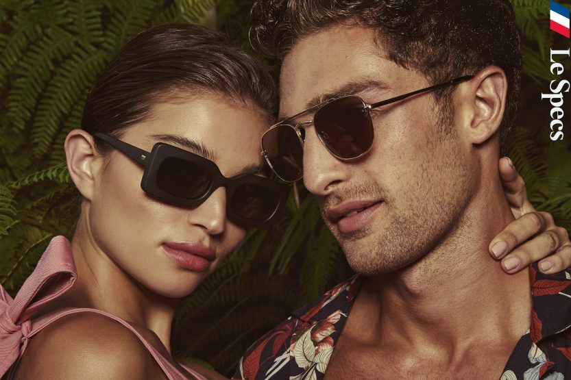 A couple poses in sunglasses against a backdrop of lush green foliage, showcasing modern eyewear and vibrant summer fashion.