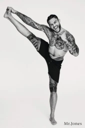 Tattooed man standing on one leg while stretching against a plain white background