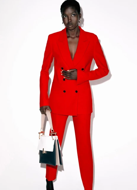Elegant Black woman in a vibrant red double-breasted suit with a stylish colour-blocked bag, standing against a clean white backdrop.