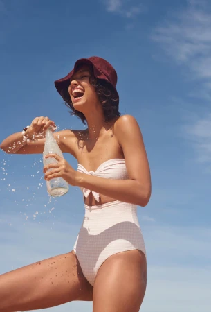 Laughing woman wearing a white swimsuit and dark bucket hat opening a bubbling glass bottle against a clear blue sky