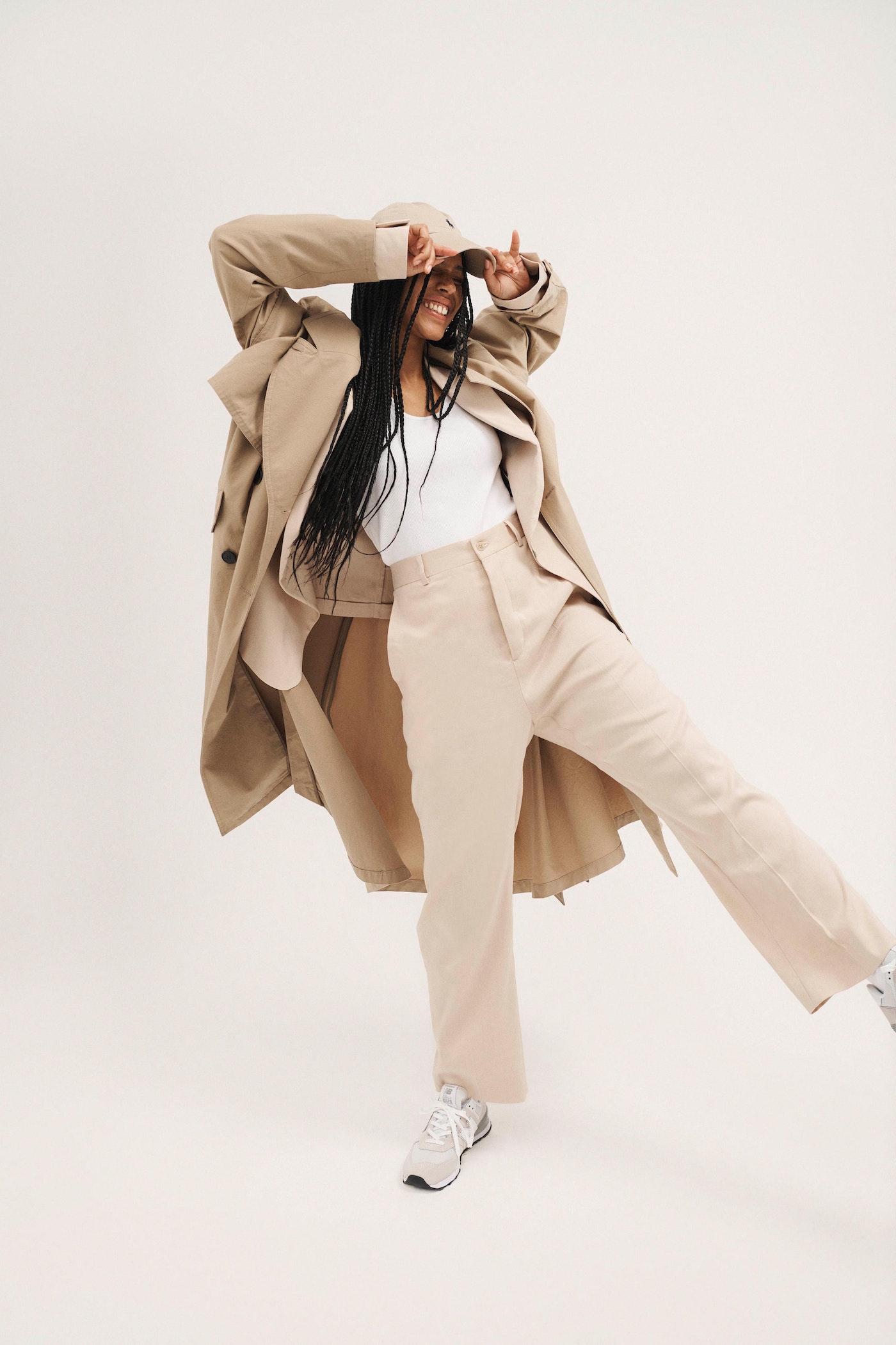 Smiling woman in a beige trench coat and cap posing against a white backdrop, showcasing modern fashion and style.