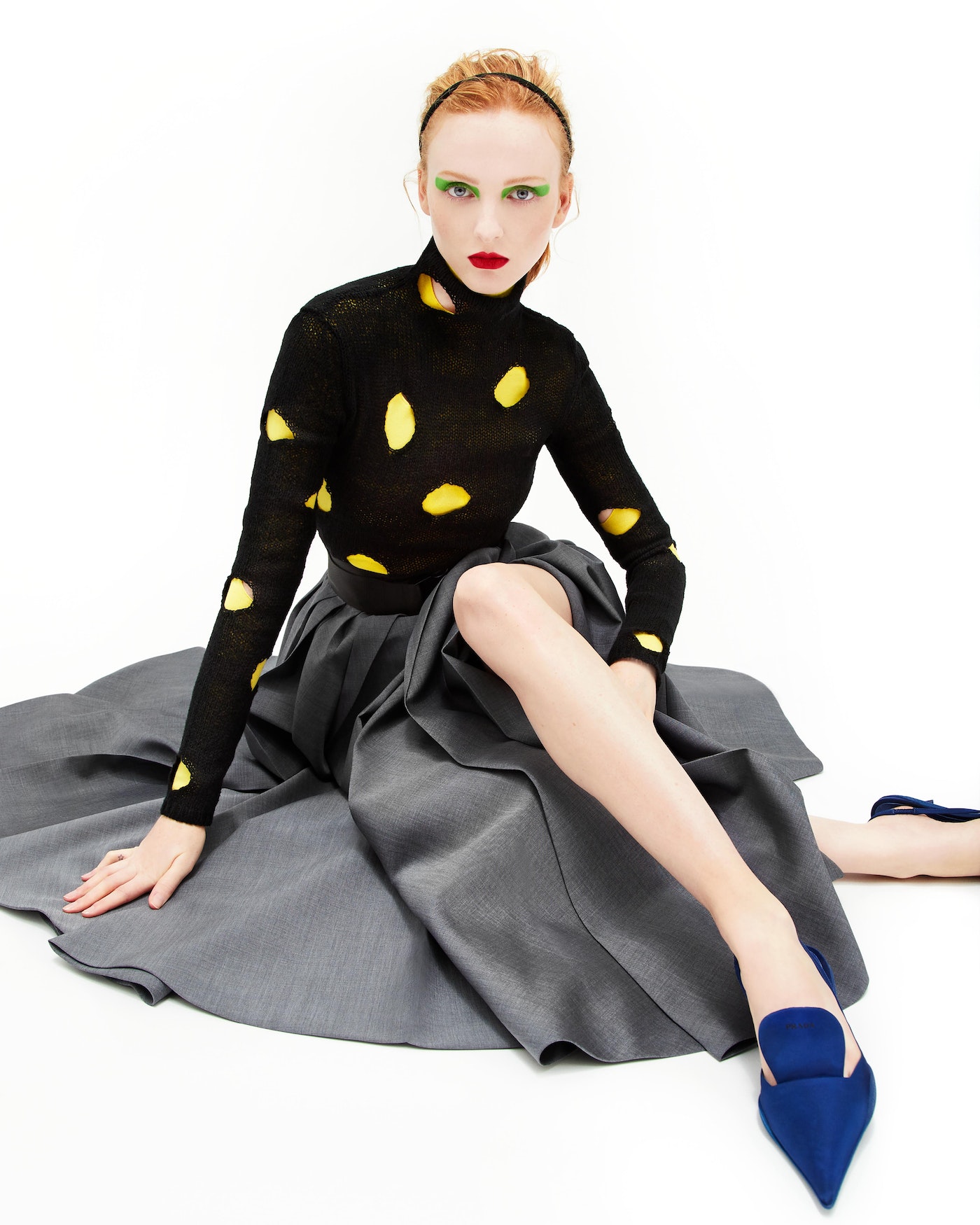 Fashionable red-haired woman in black polkadot top and grey skirt sitting against white background wearing blue heels and red lipstick.