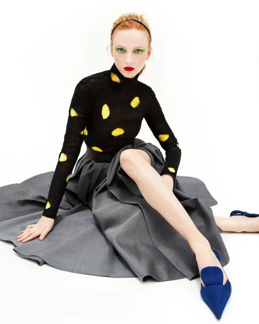 Fashionable red-haired woman in black polkadot top and grey skirt sitting against white background wearing blue heels and red lipstick.