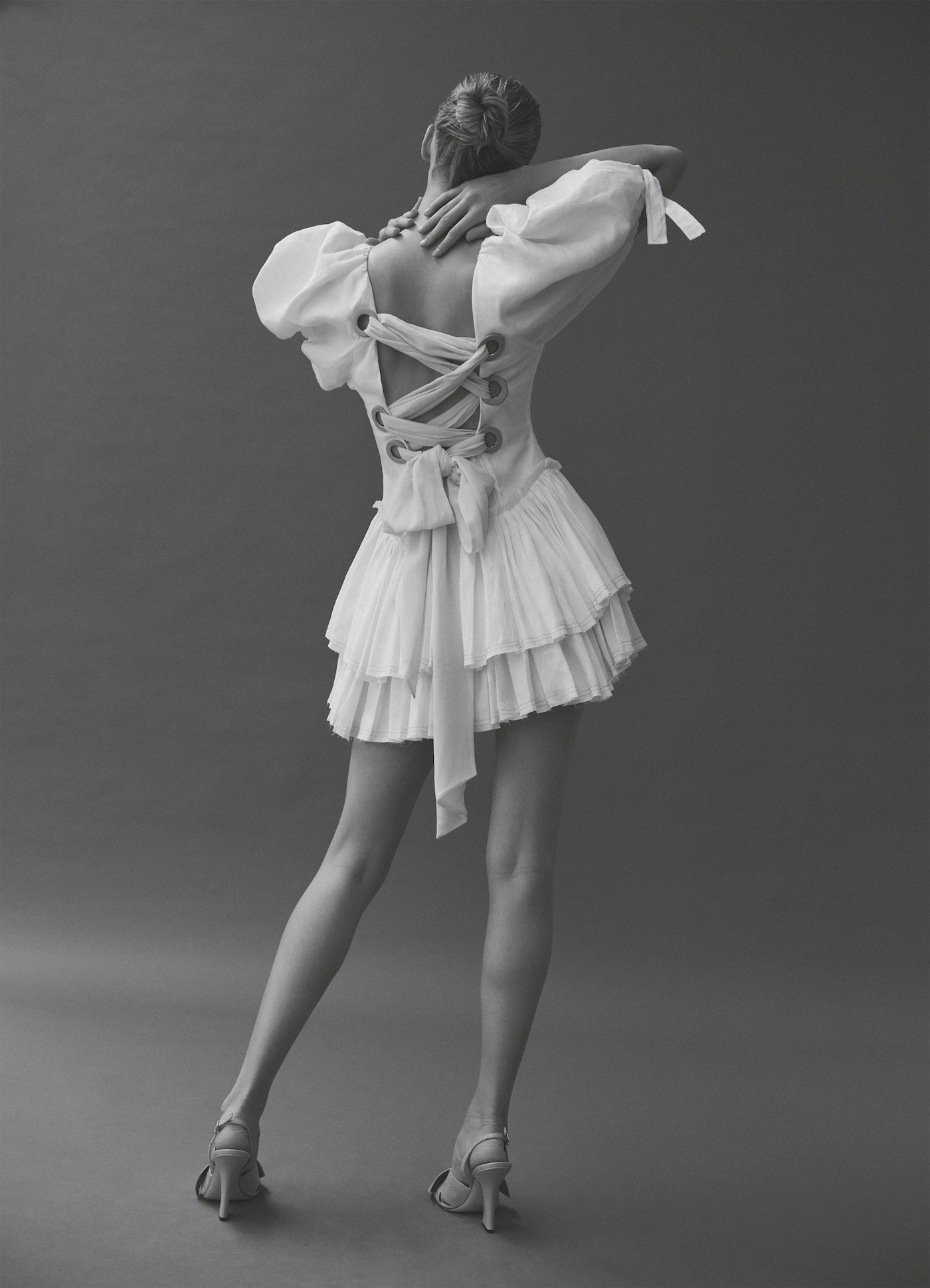 Back view of an adult woman in a white mini dress with a corset-style back, voluminous sleeves and high heels on a grey background.