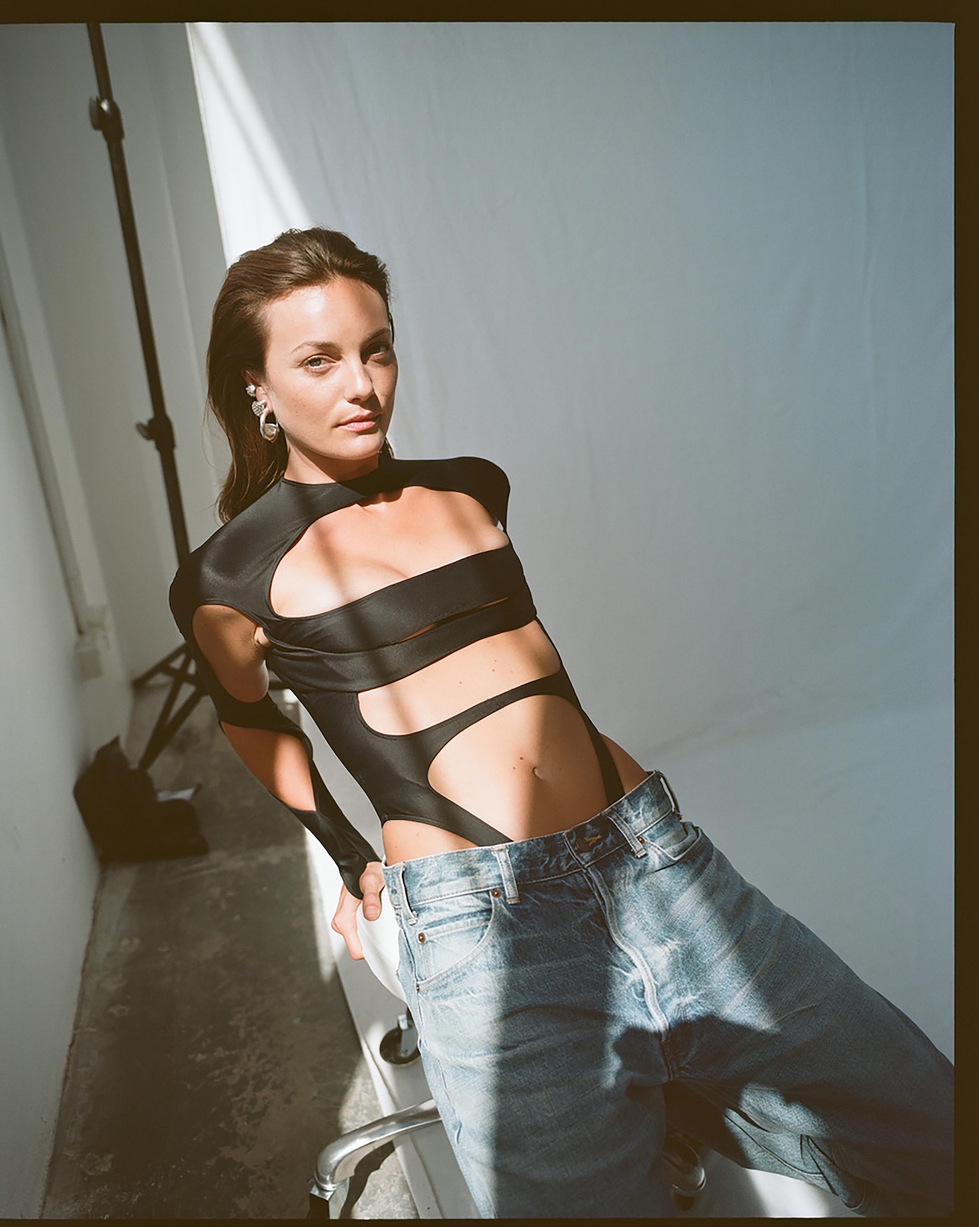 An adult woman poses in a black cut-out leotard and jeans in a photo studio with a light and stand visible against a white background.