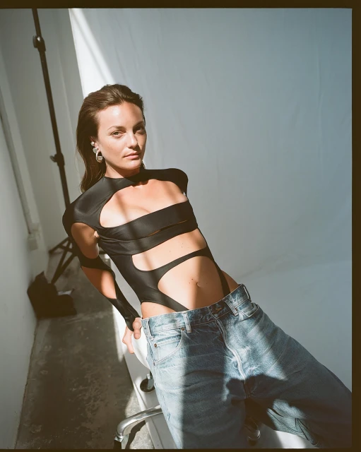 An adult woman poses in a black cut-out leotard and jeans in a photo studio with a light and stand visible against a white background.