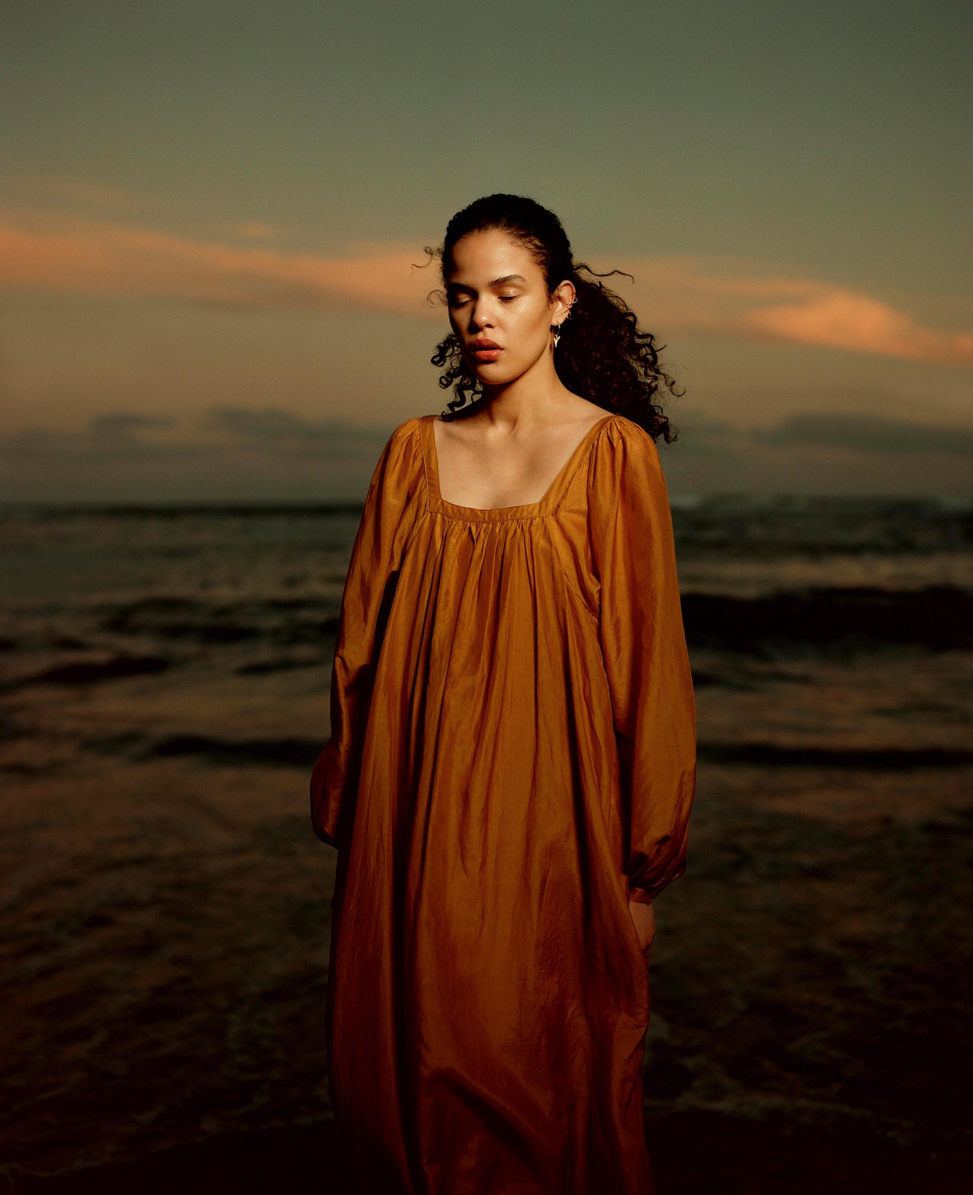 Woman in golden dress stands at the ocean's edge with a serene expression as dusk colors paint the sky above the water.