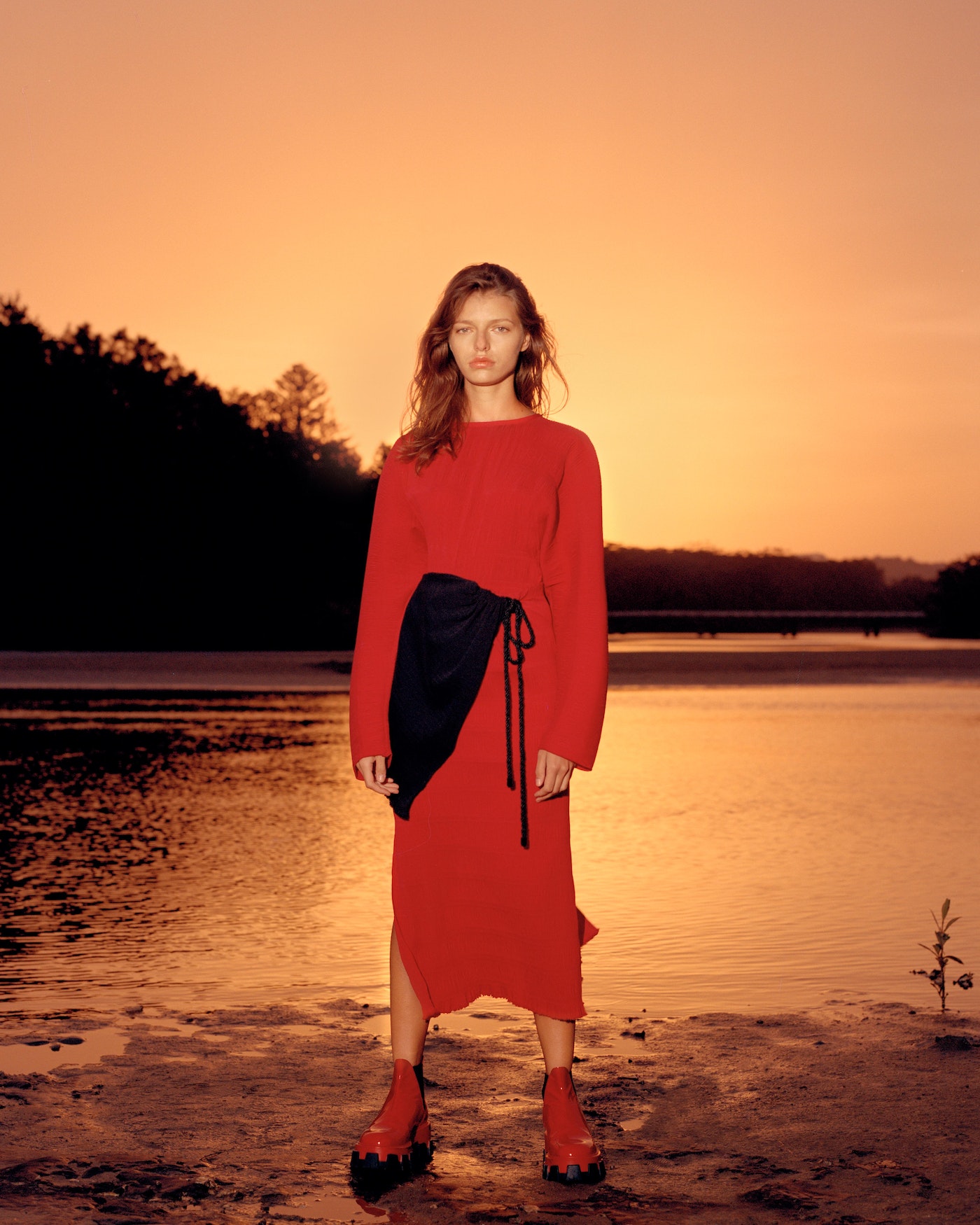 Adult woman in a red long-sleeve dress and red boots poses in a natural outdoor environment with a water backdrop and orange sky.