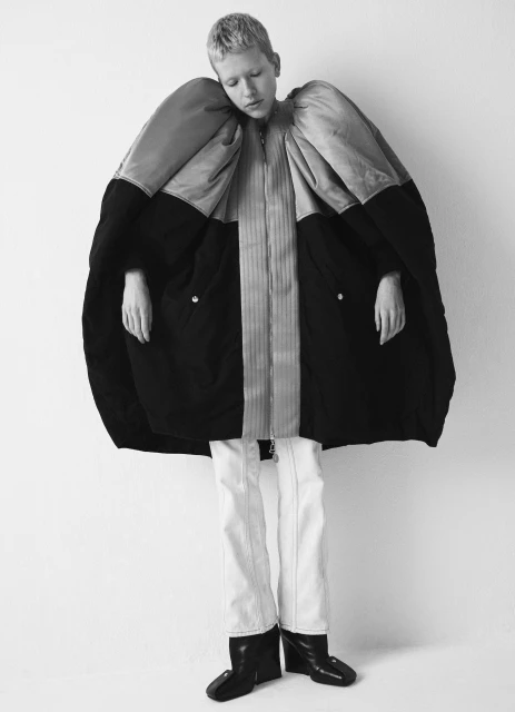 Monochrome fashion shot: androgynous model wearing oversized coat with sculptural sleeves, light pants, and wedge shoes against white background.