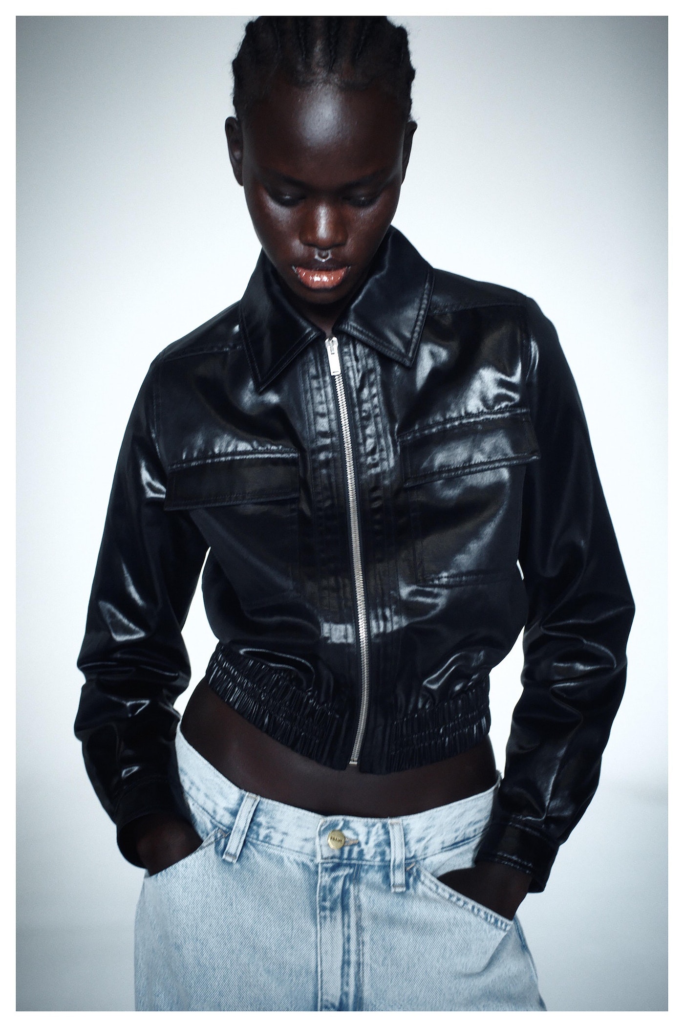 Black woman wearing a glossy black leather jacket and light wash jeans against a white seamless backdrop