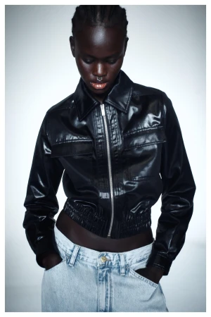 Black woman wearing a glossy black leather jacket and light wash jeans against a white seamless backdrop
