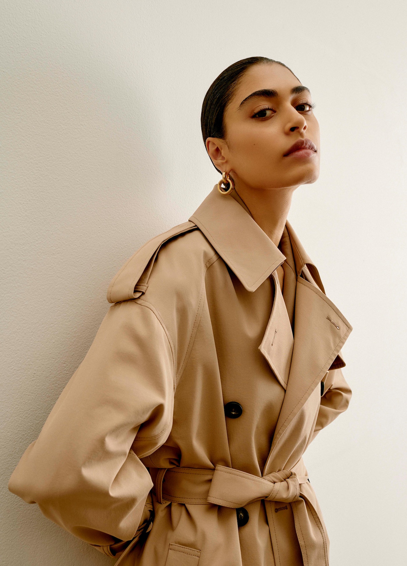 Model in camel trench coat wearing gold hoop earrings against a white wall