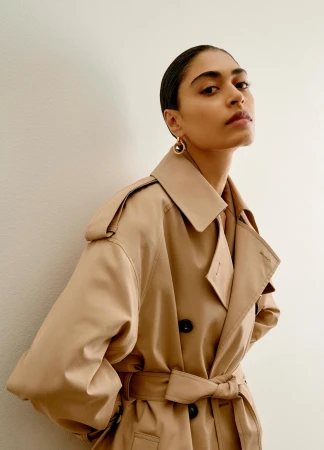 Model in camel trench coat wearing gold hoop earrings against a white wall