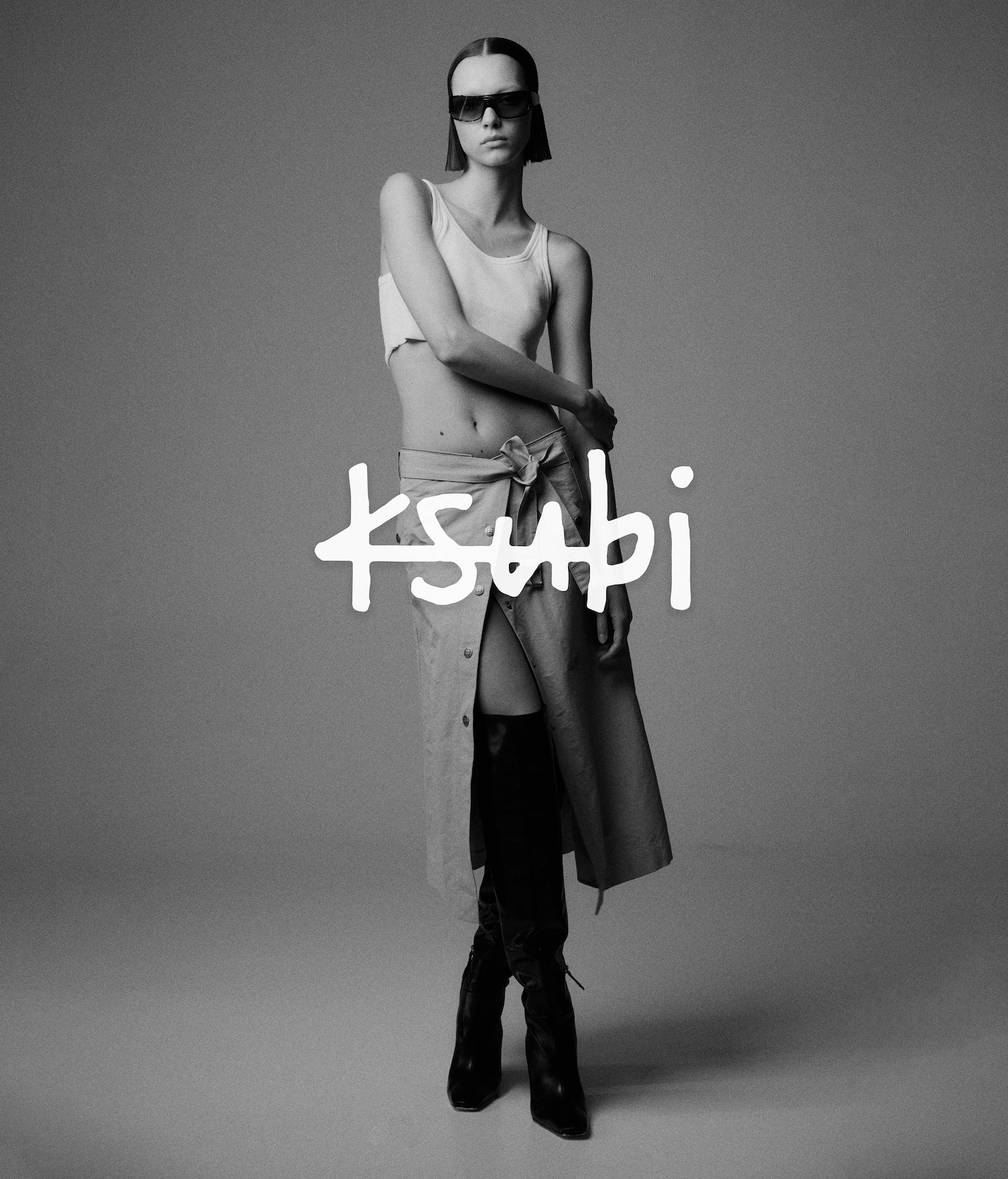 Monochrome fashion portrait of a model wearing sunglasses, white crop top, wrap skirt, and knee-high boots against a plain studio backdrop.