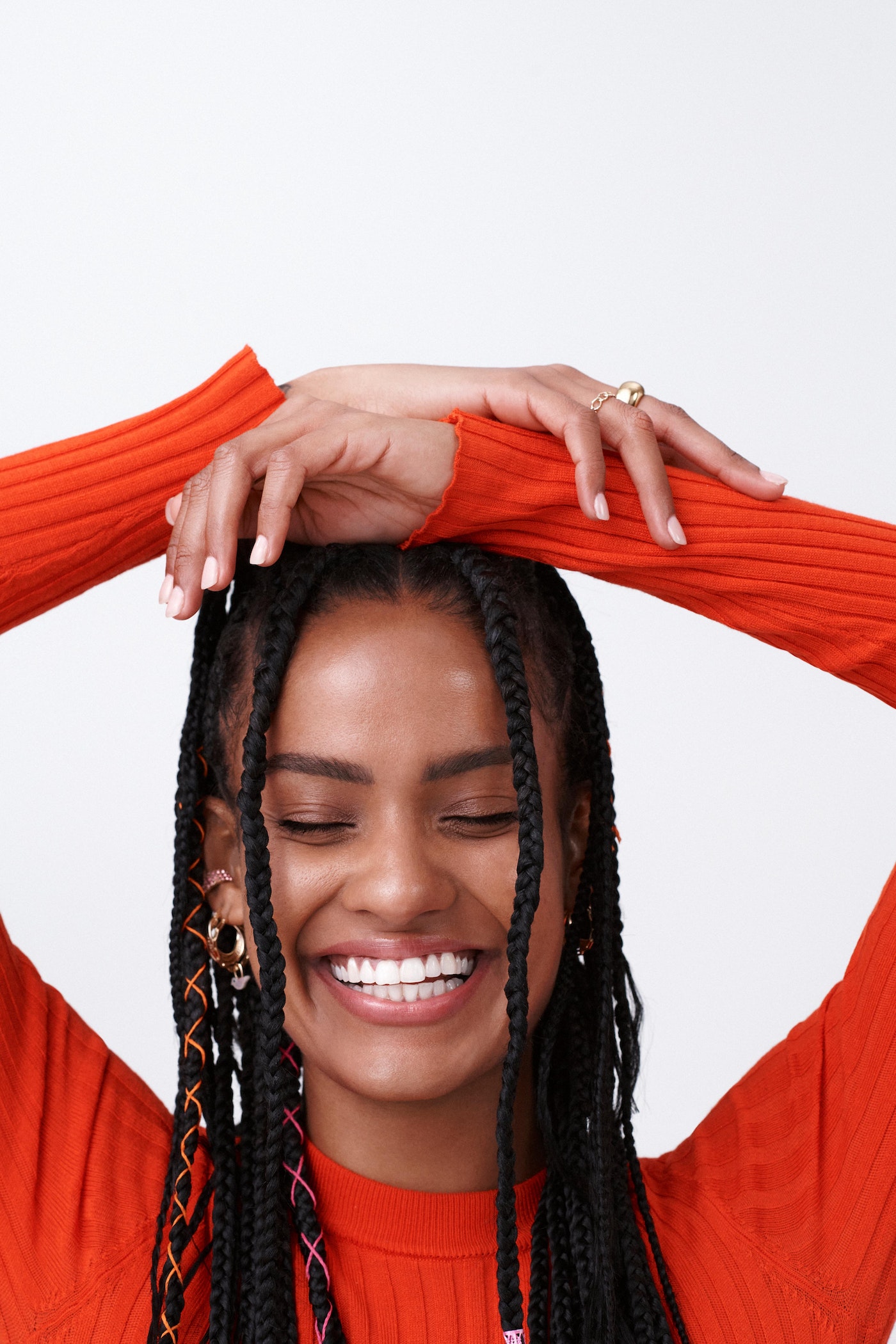 Portrait of a smiling woman with braided hair, wearing an orange long-sleeved shirt, and hands above her head on a white background.