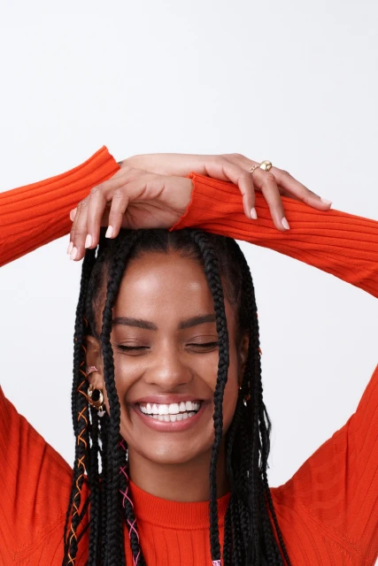 Portrait of a smiling woman with braided hair, wearing an orange long-sleeved shirt, and hands above her head on a white background.