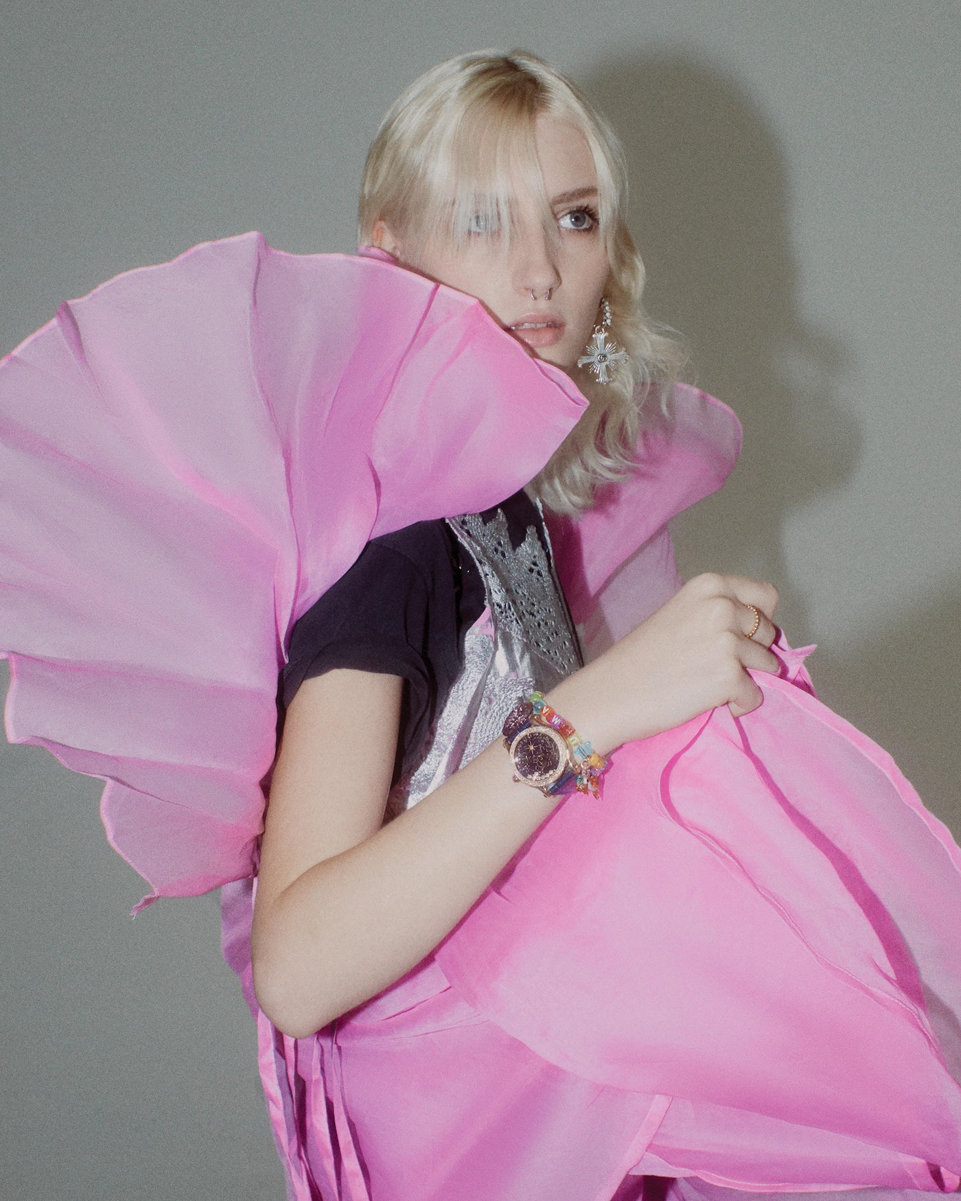 Young woman with blonde hair wearing a large pink dress and jewelry, posing in front of a grey backdrop.