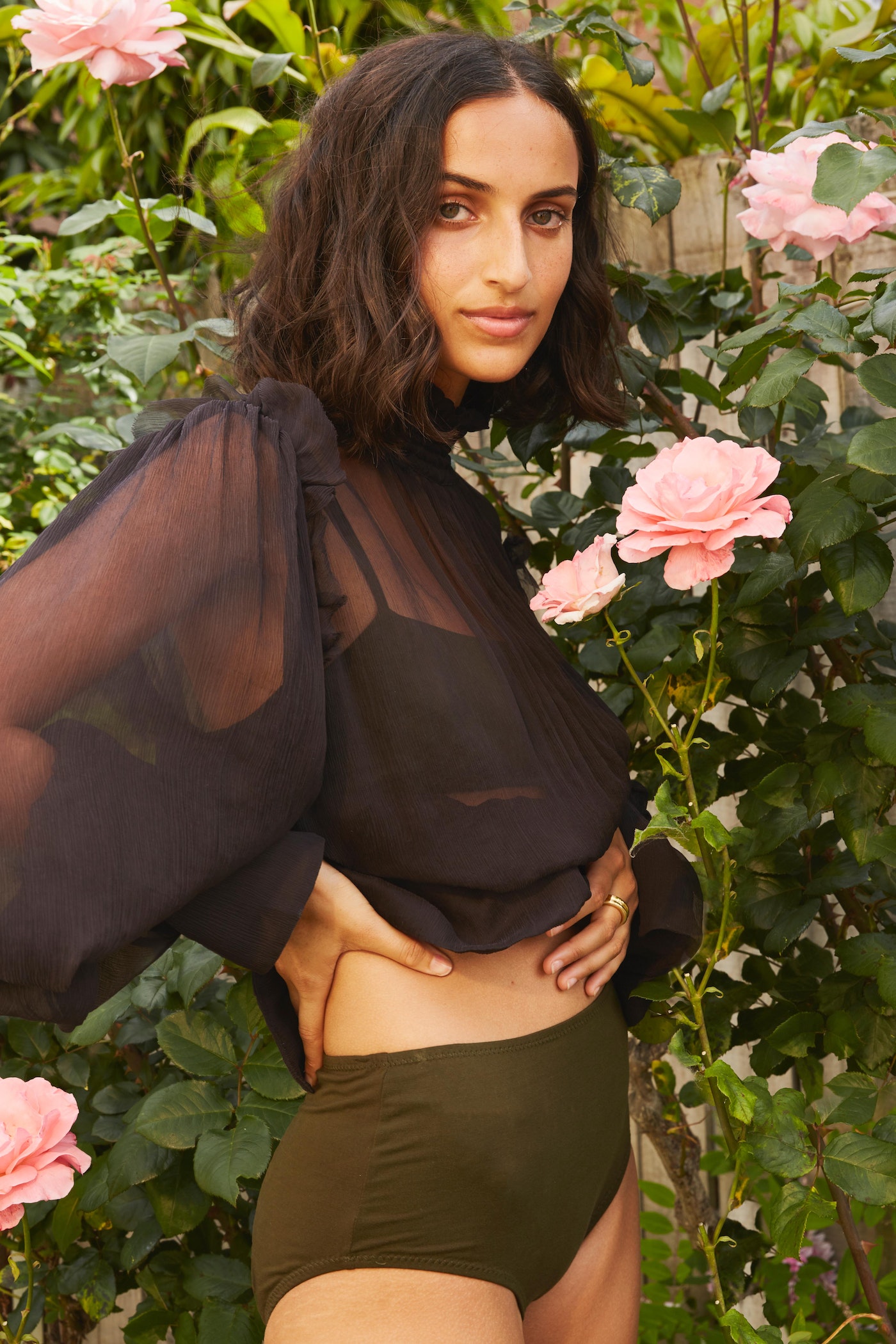 Woman in a sheer black top and olive bottoms poses outdoors amongst pink roses and green foliage in a medium shot.