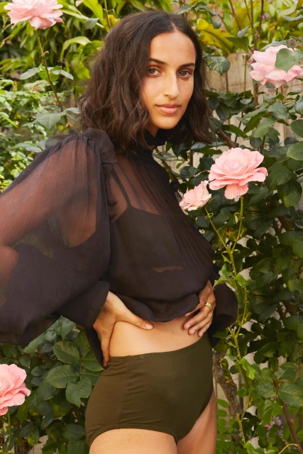 Woman in a sheer black top and olive bottoms poses outdoors amongst pink roses and green foliage in a medium shot.