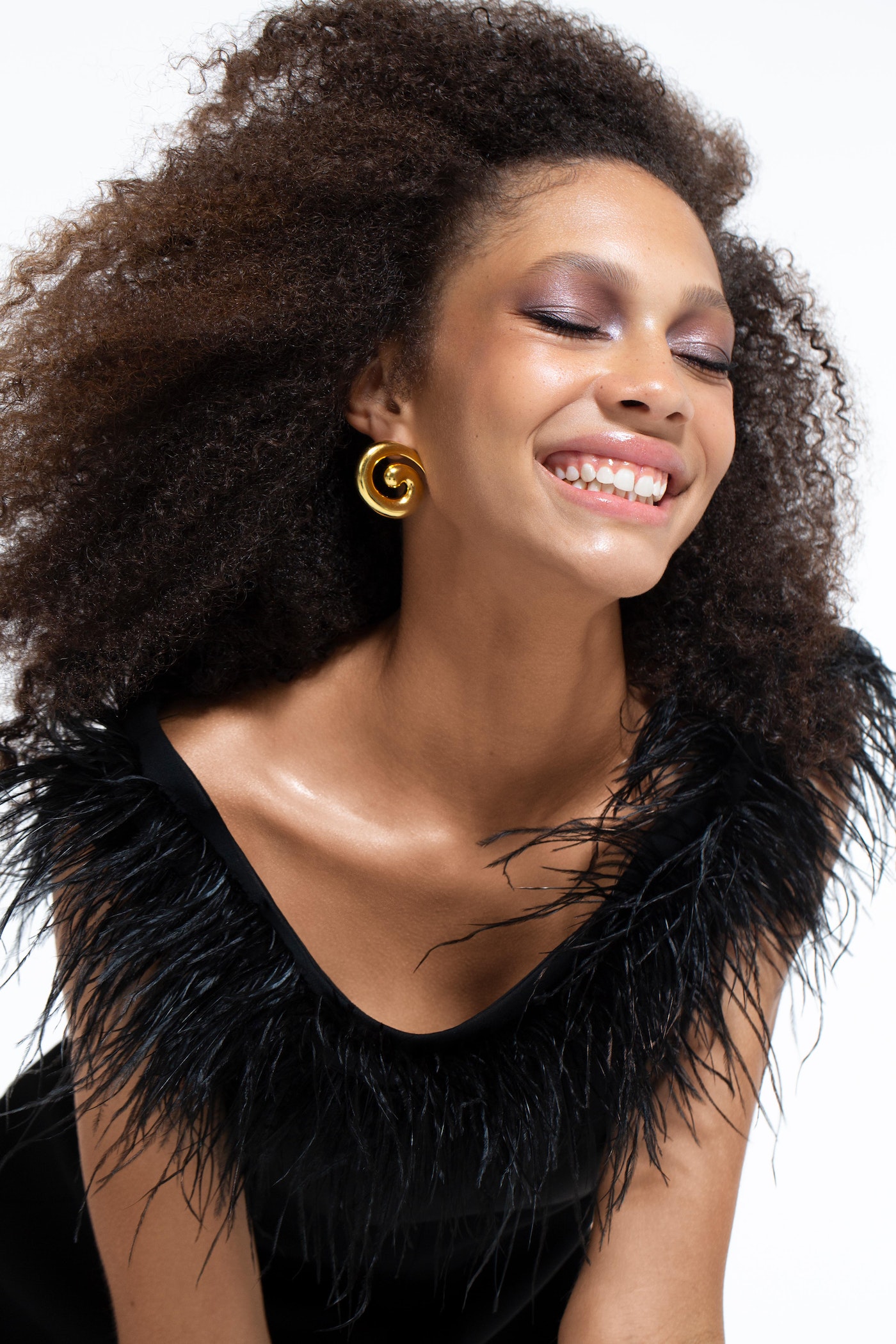 Smiling woman with curly hair and gold earrings wearing a black, feather-trimmed top against a white background in a beauty shot.