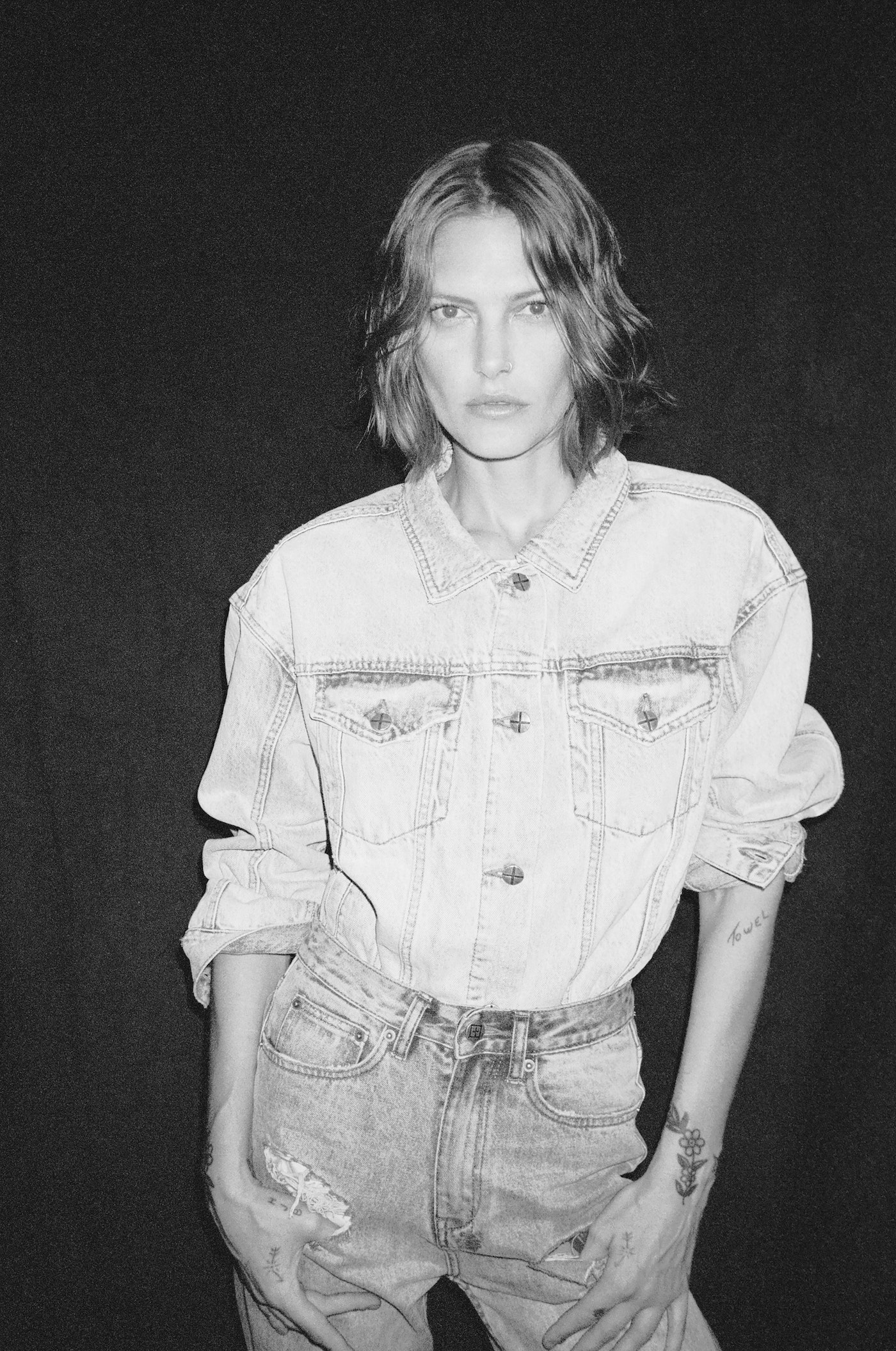 Monochrome studio portrait of a young adult woman wearing a denim jacket and jeans against a dark backdrop, fashionably styled and posed.