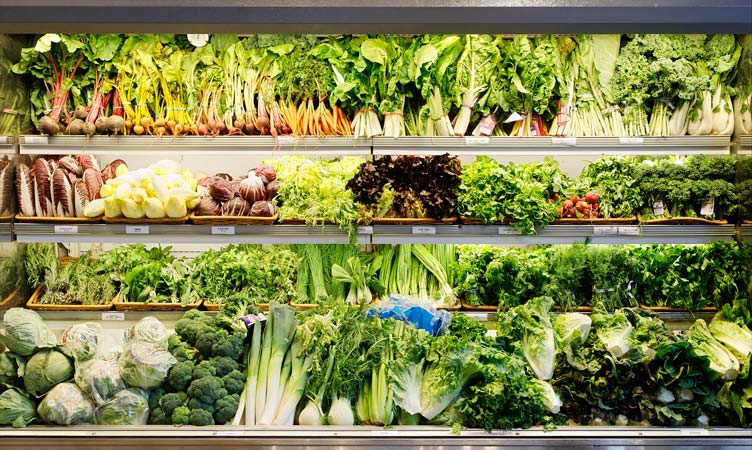 Produce aisle display featuring a variety of fresh vegetables, including leafy greens, broccoli, and root vegetables on shelves.