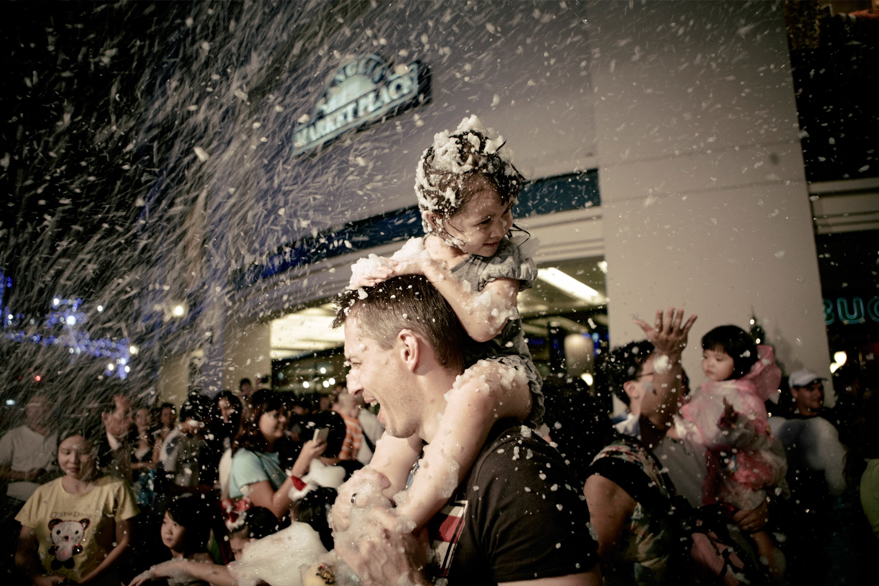 A father with his daughter on his shoulders outdoors in falling snow, surrounded by other people at night with building lights in the background.