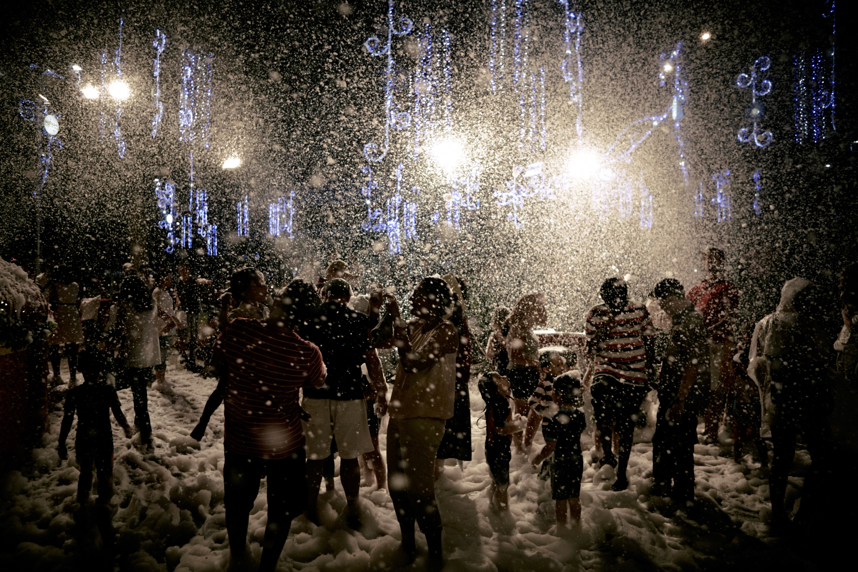 Crowd of adults and children gather outdoors at night during artificial snowfall, illuminated by string lights. Festive celebration.