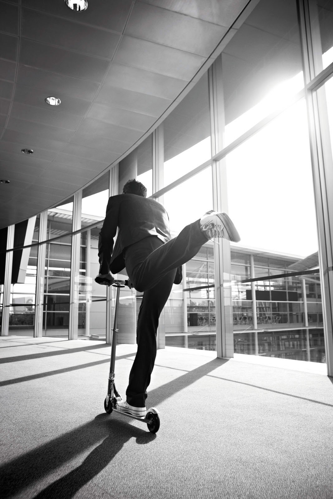 A person in a business suit balances on one leg on a scooter inside a bright, modern building with large windows, back view, in black and white.