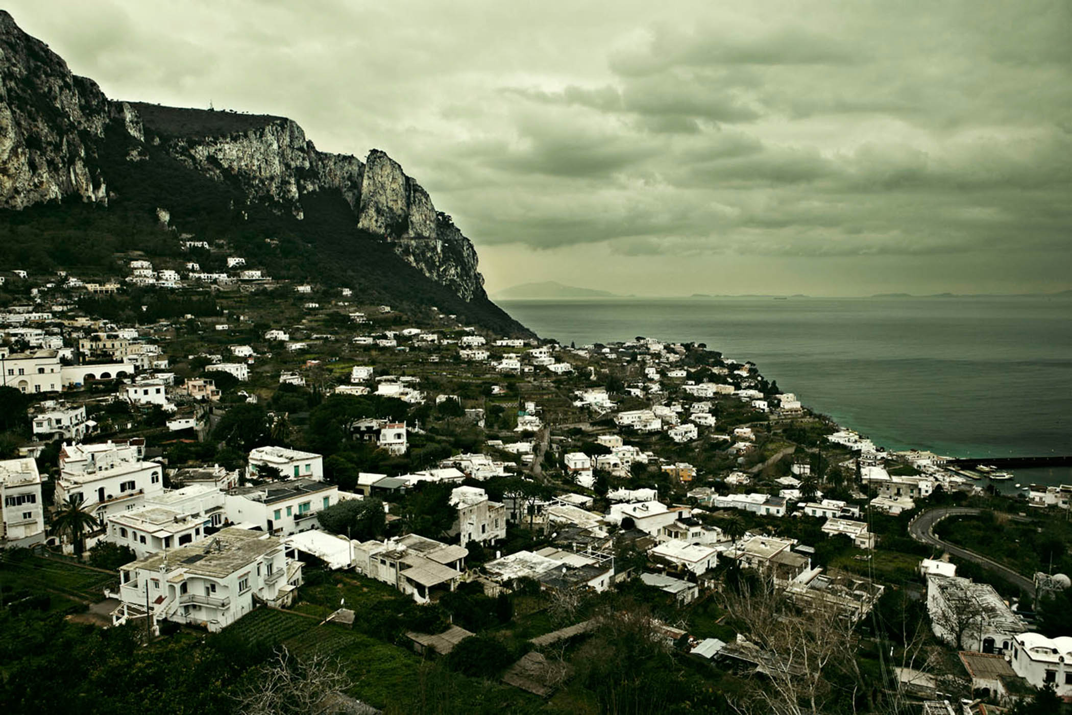 Overhead landscape view of a hillside town with white buildings under a cloudy sky, next to a body of water and a rocky mountain.