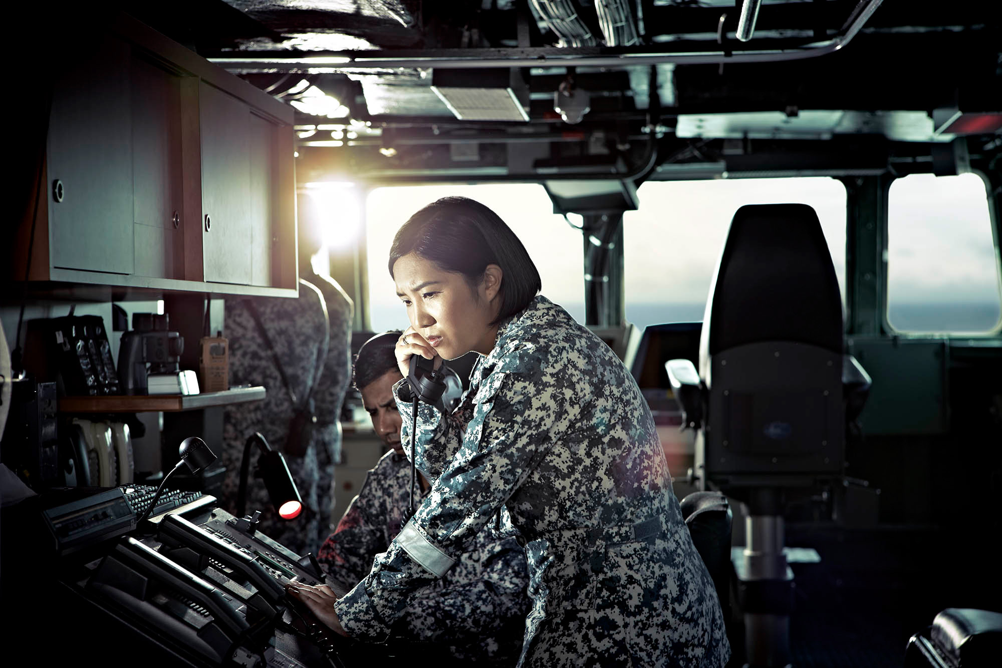 Woman in military camouflage uniform using a telephone in a ship control room, with a man nearby and navigation equipment.