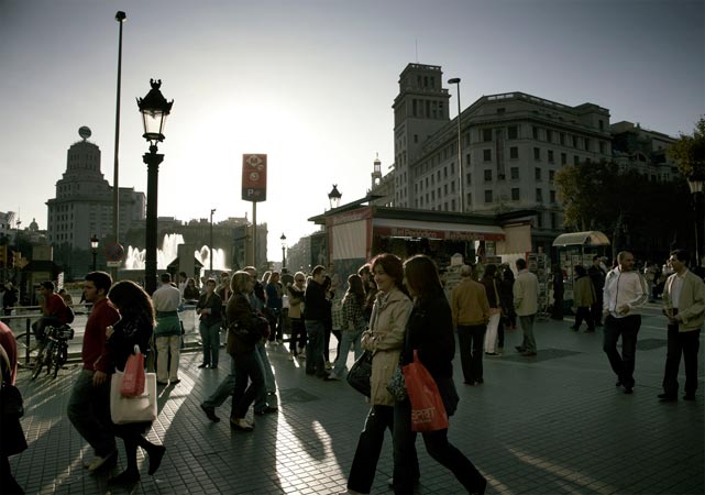 A group of pedestrians walks along a city street, surrounded by buildings, lampposts, a fountain, and trees under a bright sky.