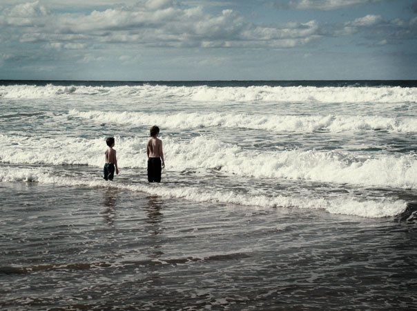 Two children stand in the shallow water at the beach, facing the ocean waves under a cloudy sky on a sunny day.