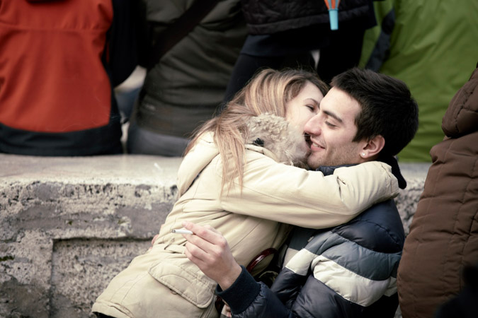 A young couple embraces and kisses outdoors, the woman holding a cigarette, with a blurred background of people and a concrete ledge.
