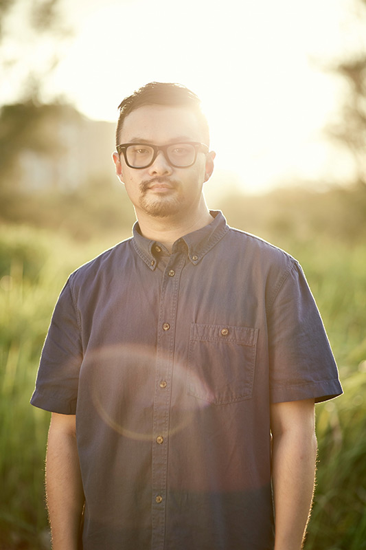 Portrait of an adult male with glasses outdoors in a field during golden hour, wearing a button-down shirt with short sleeves.