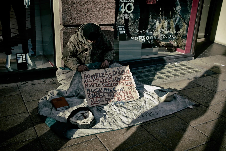 A person sitting on a pavement next to a handwritten cardboard sign and blanket outside a storefront window.