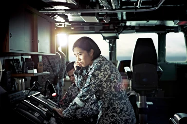 Military personnel in camouflage uniforms operating communication equipment on a ship bridge.