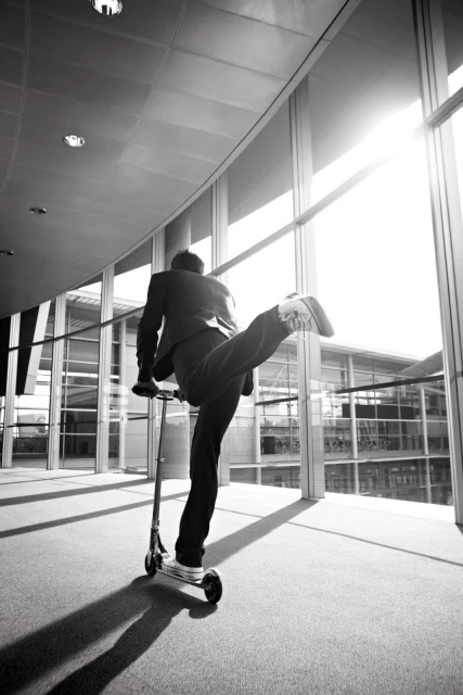 Man in a suit riding a kick scooter inside a modern building with large glass windows by Jimmy Fok