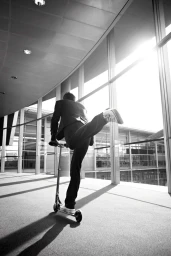 Man in a suit riding a kick scooter inside a modern building with large glass windows by Jimmy Fok