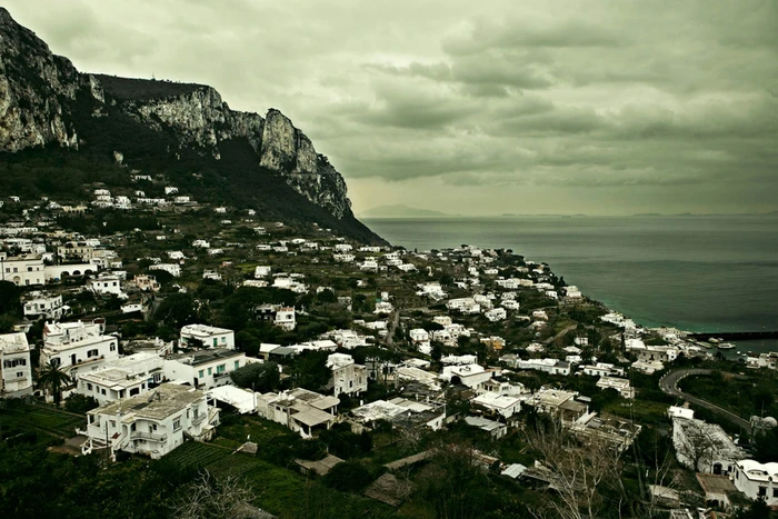Aerial view of a coastal town with white buildings nestled against steep cliffs next to a dark sea under an overcast sky
