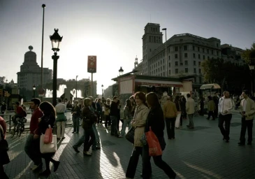 Crowded public square at dusk with people walking on paved ground and neoclassical buildings in the background.