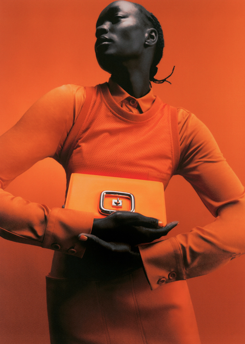 Woman in orange dress holds orange clutch purse against orange background in eye-level fashion shot with studio lighting.