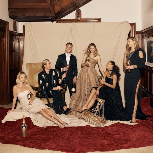 Group portrait of six fashionably dressed adults with champagne glasses, sitting and standing indoors against a tan backdrop.