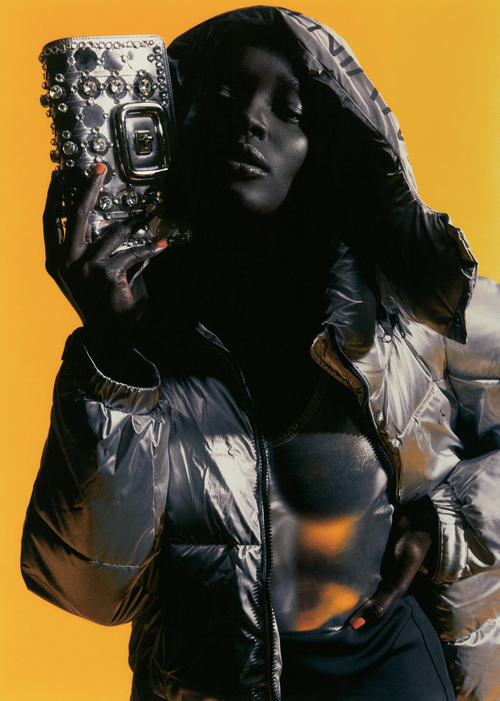 A dark-skinned model wearing a silver puffer jacket with a hood is holding a silver jeweled purse against a yellow backdrop.