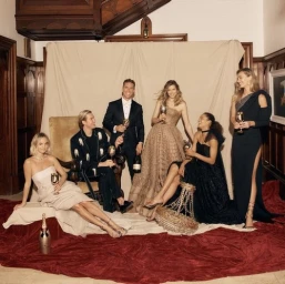 Group of six dressed-up people holding glasses in front of a neutral backdrop with a red velvet floor covering