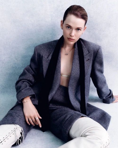 Model seated wearing a grey blazer, matching shorts, and white lace-up boots against a textured grey studio background.