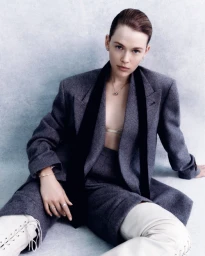 Model seated wearing a grey blazer, matching shorts, and white lace-up boots against a textured grey studio background.