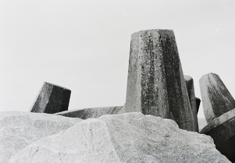 Monochrome image features concrete tetrapods on a rock, under an overcast sky, providing a stark, minimalist seascape scene.
