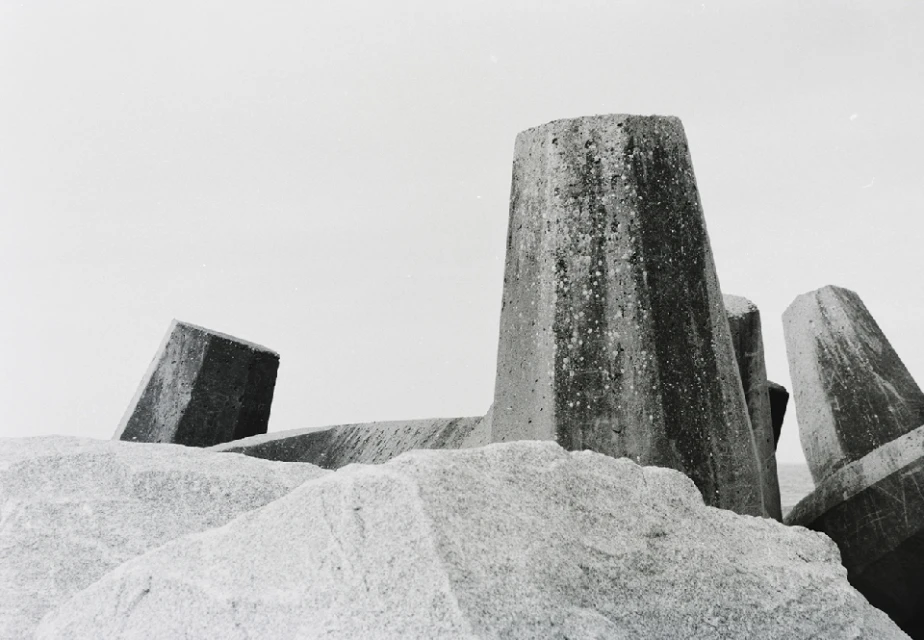 Monochrome image features concrete tetrapods on a rock, under an overcast sky, providing a stark, minimalist seascape scene.